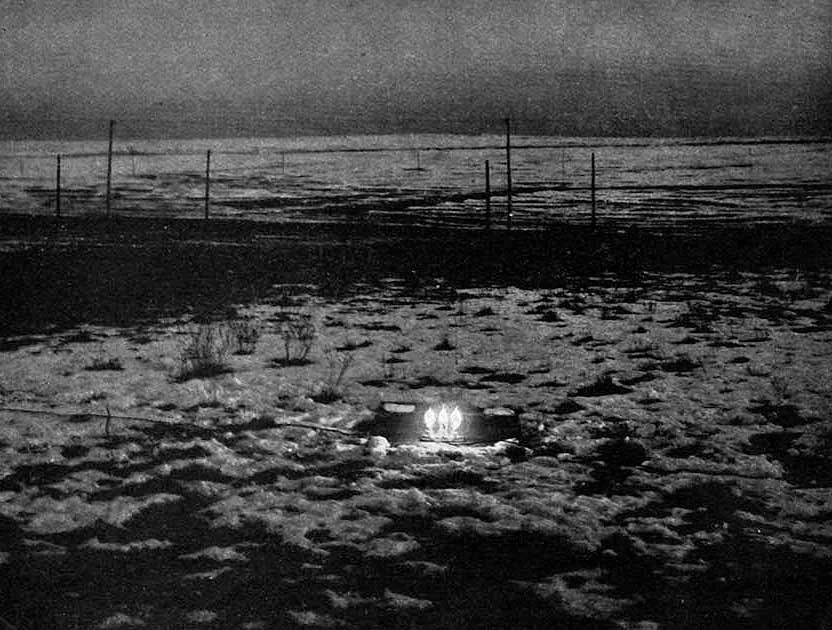 A dark night photograph looking across flat scrubby open ground. In the middle distance, close to the ground, three incandescent lamps burn brightly in a small cluster, the only light in the frame, with a thin wire leading away to the left. A line of fence posts and wire crosses the middle of the picture, and the horizon is a faint band at the top.