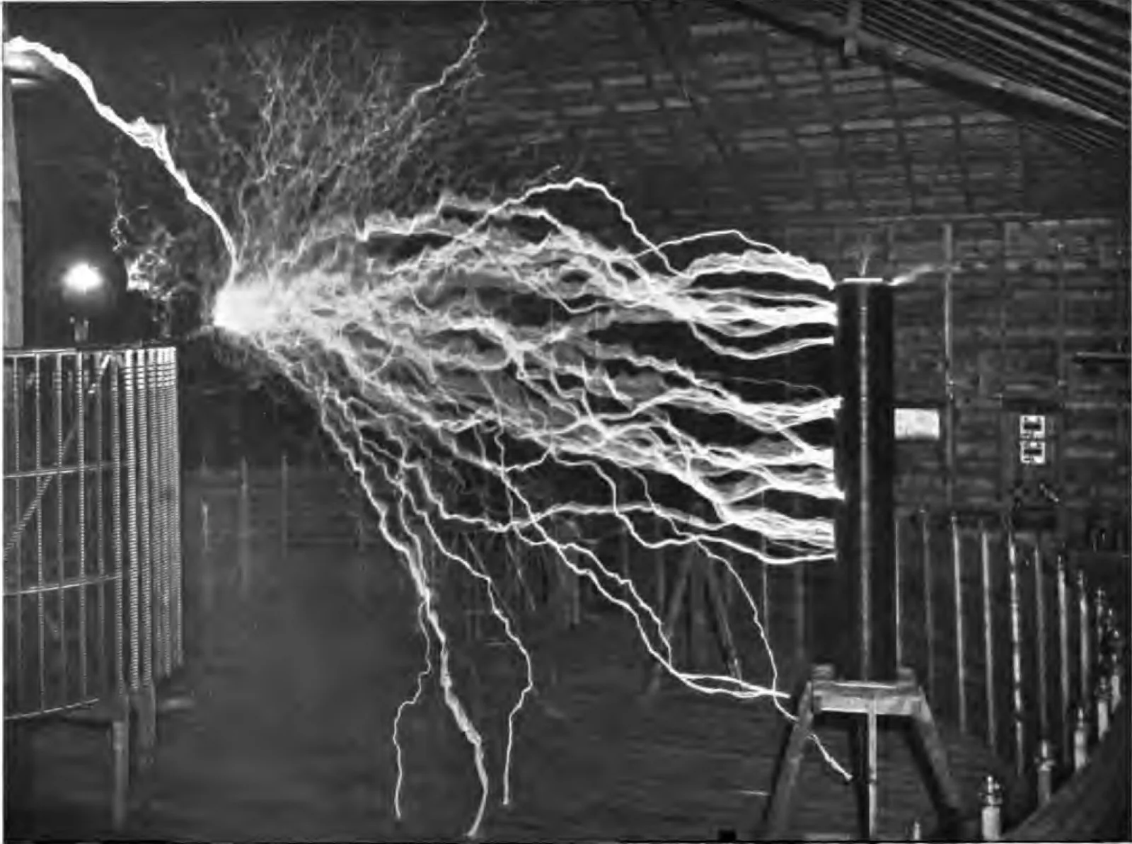 A black and white photograph of the inside of a wooden barn at night. A dense fan of white branching electrical discharge erupts from a point at the upper left and sprays across most of the frame to the right, striking a dark cylindrical coil standing on a low wooden trestle. A tall cage-like coil wound with closely spaced wire stands at the left edge. Bare plank walls and open roof timbers are visible behind. There is no chair and no person in the picture.