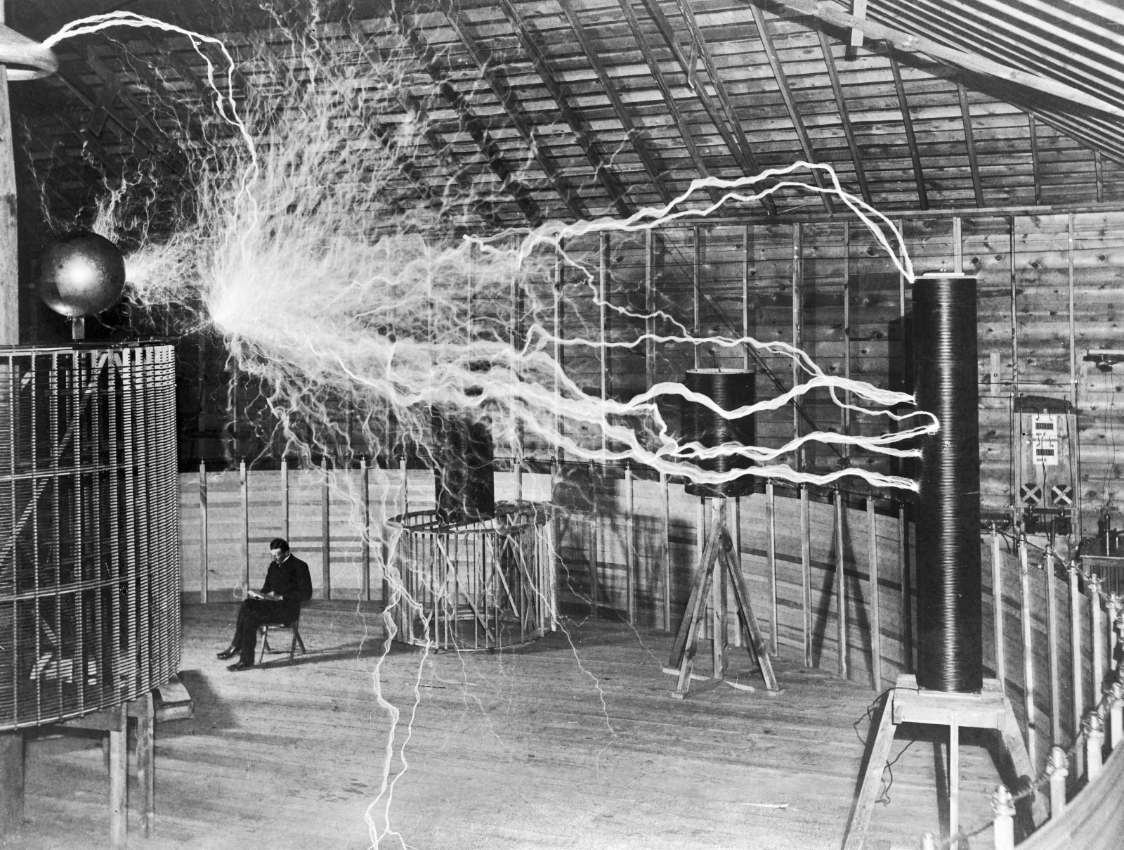 Photograph of a large timber-framed shed. A huge circular coil fills the left of the frame and a tall cylindrical coil stands at the right. Ragged white discharges many feet long spray from a metal sphere at the top left and arc across the whole width of the room. A man in a dark suit sits calmly on a folding chair in the middle of the floor, reading a book, directly beneath the discharge, apparently untouched by it.