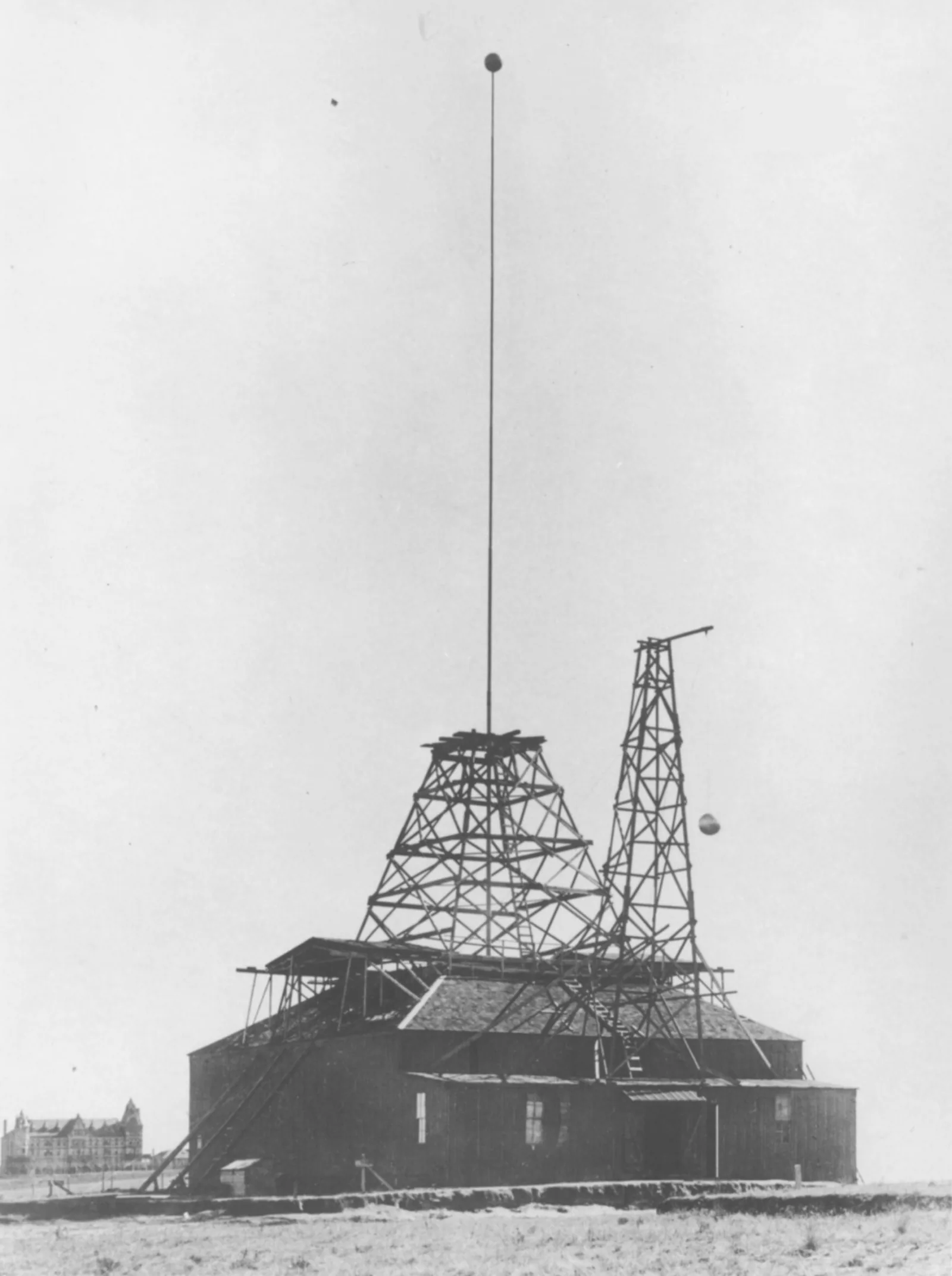 A black and white photograph of a low wooden barn standing alone on flat bare prairie. A pyramidal timber framework rises out of the middle of its roof, and from the top of that framework a very tall, very thin mast climbs most of the height of the picture, with a small dark ball at its tip. A second, narrower lattice derrick stands beside it with a sphere hanging from a projecting arm. A wooden stair runs up the left-hand end of the building. A large institutional building sits on the horizon at far left.