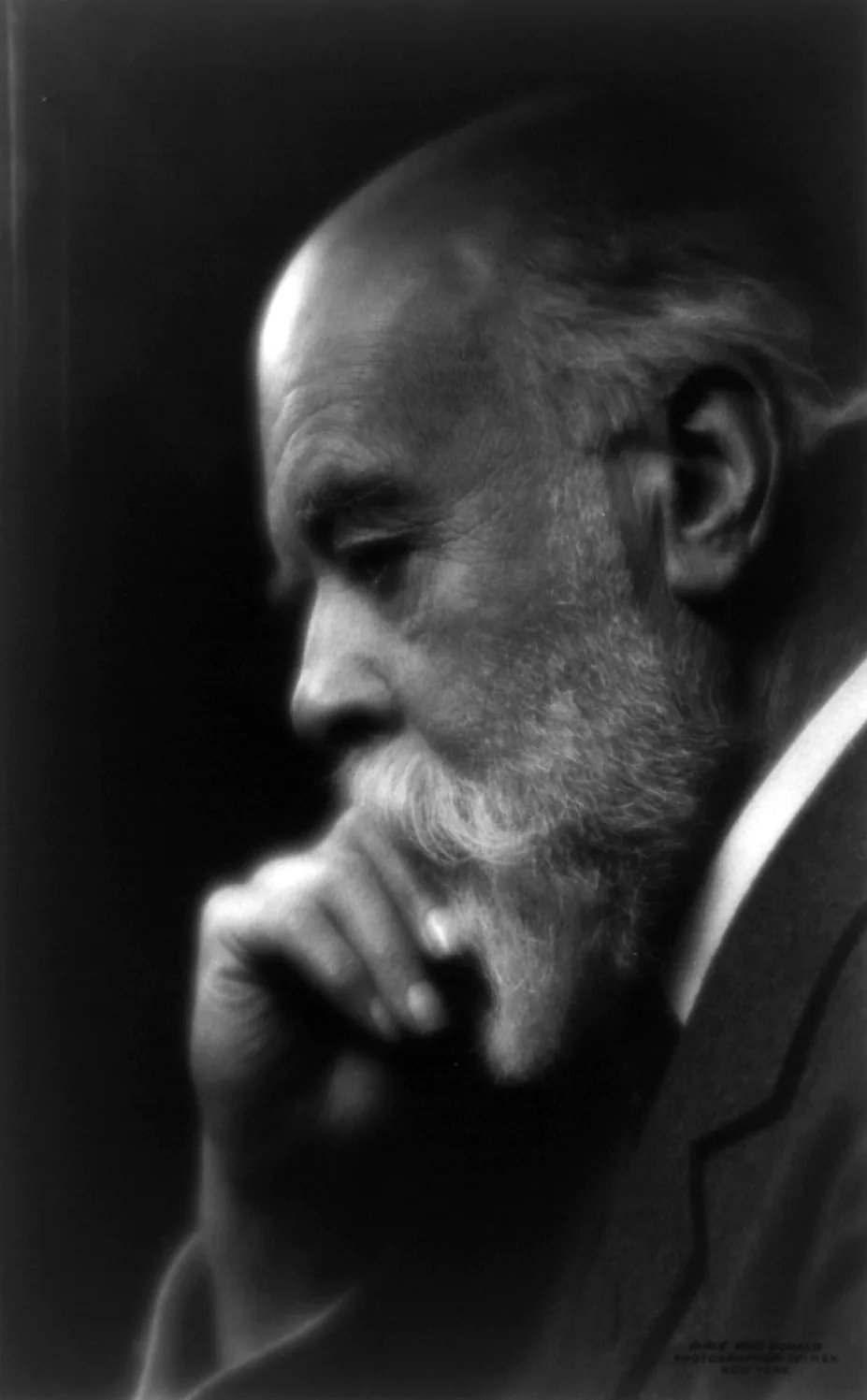 Studio photograph of an elderly bearded man in near profile against a dark background, lit from the left. He is bald on top with white hair at the sides, has a full white beard and moustache, and rests his chin on the knuckles of one hand. He wears a dark jacket with a white collar and looks downward, away from the camera.