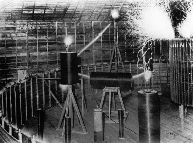 Photograph inside a wooden shed with a plank floor and an open-raftered roof. Several dark cylindrical coils stand on rough timber trestles of different heights. Brilliant white discharges stream from metal balls mounted above two of the coils and from a curved rail at the right, lighting the boards behind them. The room is otherwise empty.