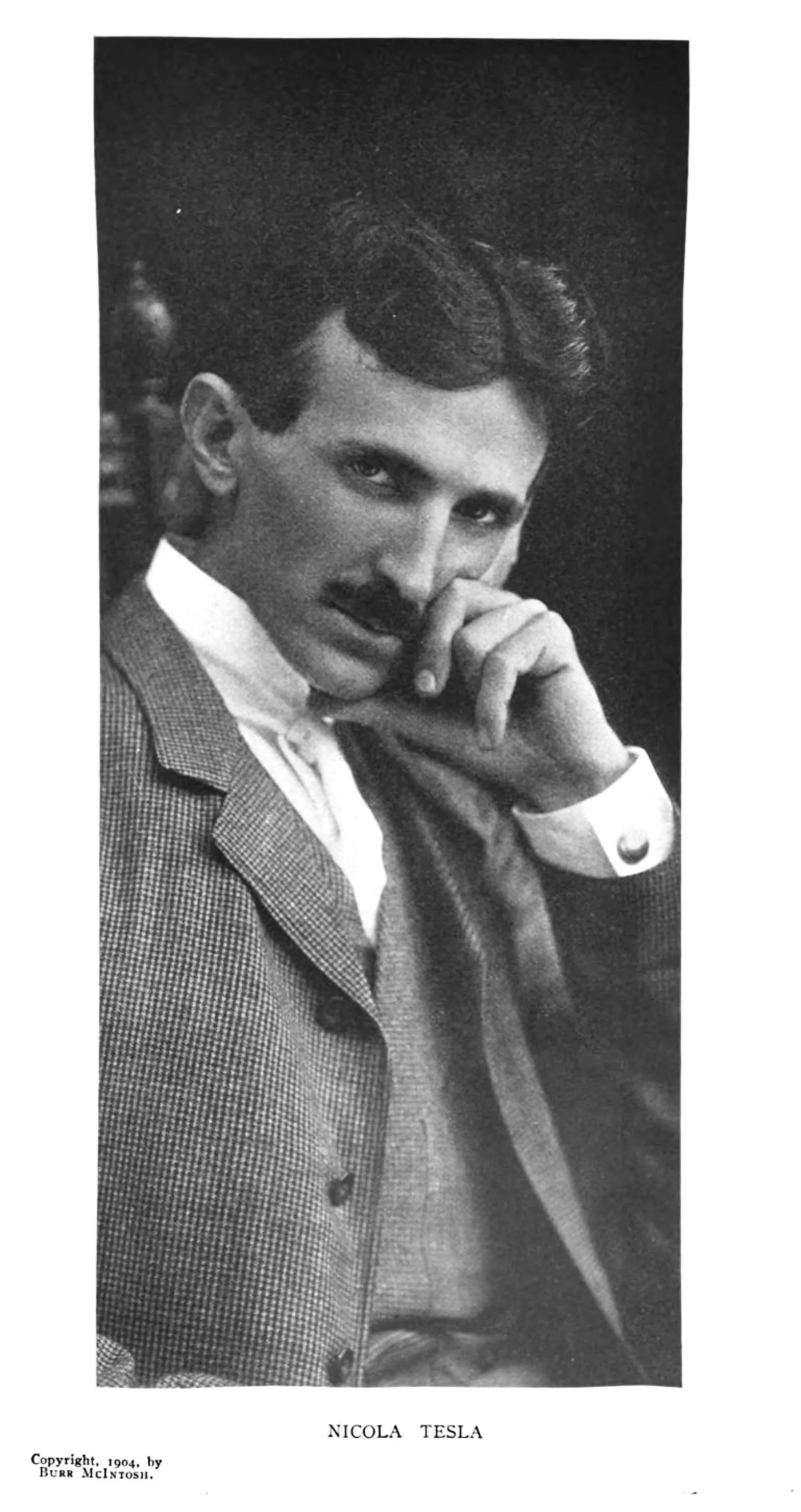 A magazine page carrying a tall studio photograph of a man in his forties. He wears a light checked jacket over a high white collar and a dark cravat, and rests his chin against the knuckles of one hand, looking straight at the camera. Printed beneath the picture is the caption NICOLA TESLA, and in the lower left corner of the page, in small type, the words Copyright 1904 by Burr McIntosh.