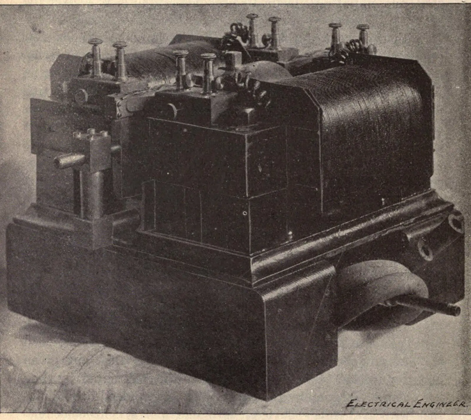 A sepia halftone photograph of a machine standing alone on a plain studio floor. It is a heavy cast-metal assembly, roughly cubical and wider than it is tall, resting on a broad moulded bedplate. A large cylindrical coil wound with dark wire fills the right half; valve stems with knurled caps project upward along the top; a short rod and a small pillar stand at the left, and a tapered air pipe enters at the lower right. The words ELECTRICAL ENGINEER are lettered into the lower right corner of the plate.