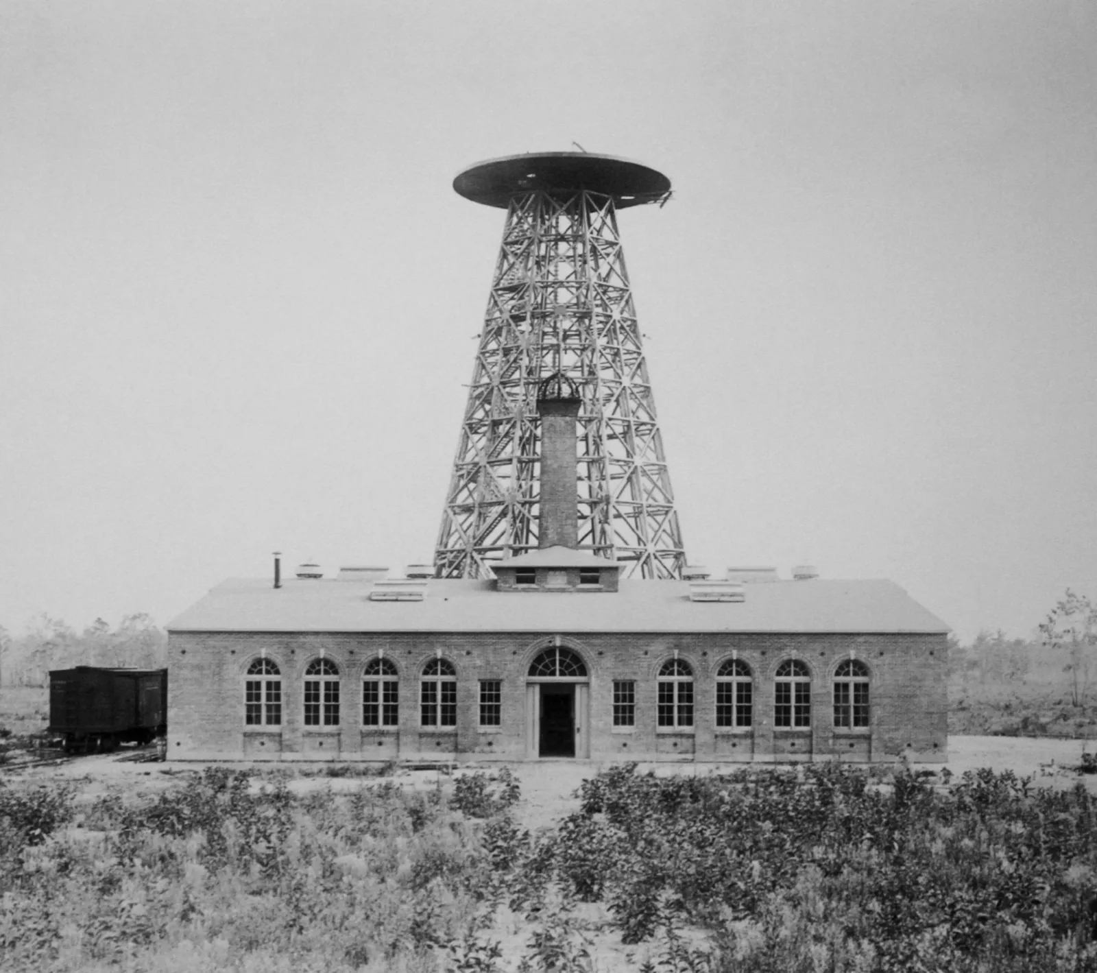 Black and white photograph of a wide, low brick building standing alone in scrub. Rising from behind its roof ridge is a tapering octagonal lattice tower of crossed timbers, topped by a broad flat disc. A square brick chimney climbs through the middle of the tower's legs, ending just below the disc. A single railway boxcar stands on a siding at the left; the ground in front is rough with low brush.